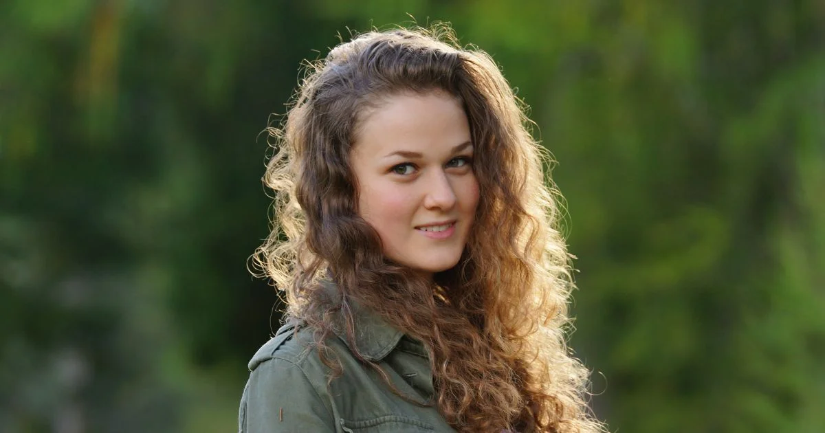 Shari Ann Chinnis Indianapolis, a young woman with curly hair and a warm smile, standing outdoors in a green jacket, with a blurred, lush green background.